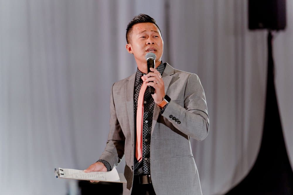 Professional bilingual MC in gray suit speaking confidently into wireless microphone at wedding reception