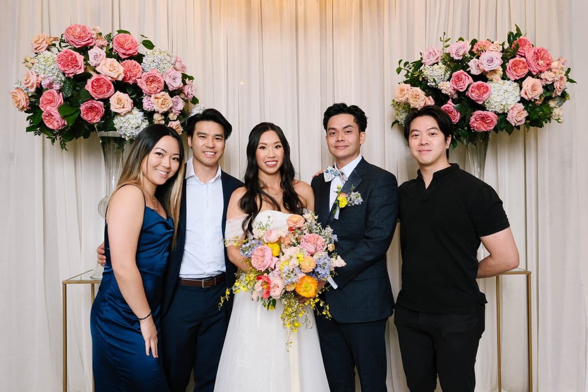 Bride with white bouquet and groom with wedding party against elegant curtain backdrop with pink floral arrangements